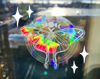 May include: A holographic sticker featuring a white outline of a butterfly with a rainbow iridescent background. The sticker is on a window with a blurry background of a city and mountains.