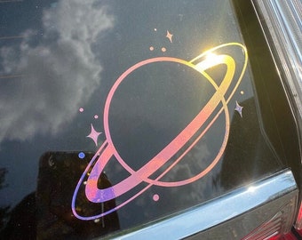 Könnte beinhalten: Ein holografischer Aufkleber eines Planeten mit Ringen auf einem schwarzen Autofenster. Der Aufkleber ist schillernd in Rosa-, Lila- und Blautönen.