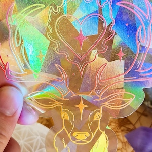 May include: A holographic sticker featuring a deer design with antlers, a heart, and geometric shapes. The sticker is held in front of a book titled "Queen of Shadows" by Sarah J. Maas. Other items include crystals and a wooden coaster.