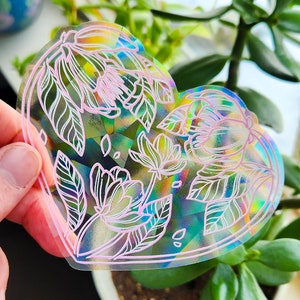 Op de afbeelding: Een hartvormige, iriserende sticker met een wit bloemenontwerp en roze contouren. De sticker wordt in een hand gehouden tegen een groene plantenachtergrond.