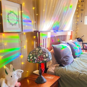 May include: A bedroom with a bed, a nightstand, a lamp, and a stuffed animal. The bed has a white canopy and a green duvet. The nightstand is made of wood and has a drawer. The lamp is a stained glass lamp with a floral design. The stuffed animal is a white bunny with wings.