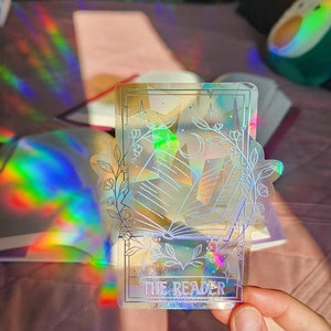 May include: A holographic sticker with a white outline of a book, a crescent moon, and stars. The sticker has the text "THE READER" at the bottom.