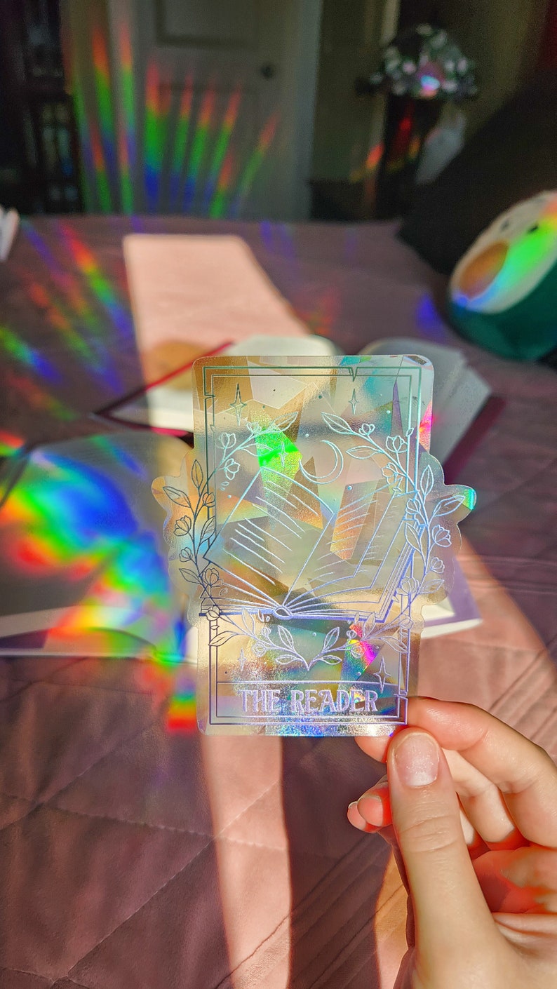 May include: A holographic sticker with a white outline of a book, a crescent moon, and stars. The sticker has the text "THE READER" at the bottom.