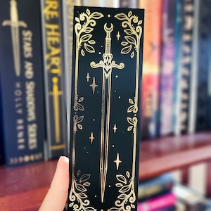 Può includere: Un segnalibro nero con un designo in foglia d'oro di una spada con una luna crescente e stelle. La spada è circondata da foglie e viti in foglia d'oro.