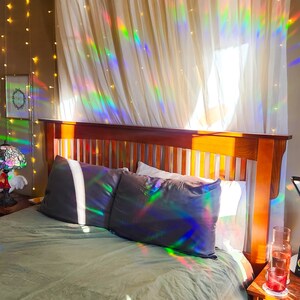 May include: A bedroom with a wooden bed frame and a green duvet. The bed is made with two pillows and a white sheet. The curtains are white and sheer, with a rainbow prism effect from the sunlight shining through. There are string lights hanging on the wall behind the bed.