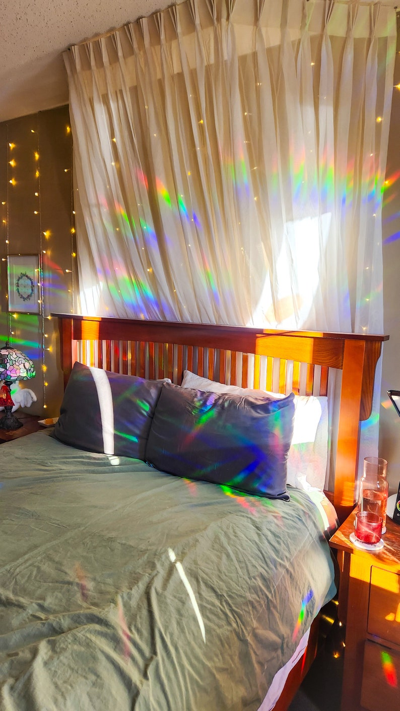 May include: A bedroom with a wooden bed frame and a green duvet. The bed is made with two pillows and a white sheet. The curtains are white and sheer, with a rainbow prism effect from the sunlight shining through. There are string lights hanging on the wall behind the bed.