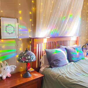 May include: A bedroom with a bed, a nightstand, and a lamp. The bed has a green duvet and two pillows. The nightstand is made of wood and has two drawers. The lamp is a stained glass lamp with a floral design. The walls are beige and have string lights hanging on them. There is a sheer curtain hanging over the bed.