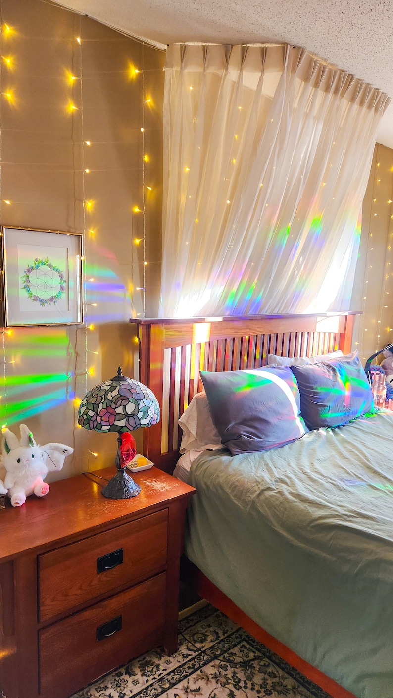May include: A bedroom with a bed, a nightstand, and a lamp. The bed has a green duvet and two pillows. The nightstand is made of wood and has two drawers. The lamp is a stained glass lamp with a floral design. The walls are beige and have string lights hanging on them. There is a sheer curtain hanging over the bed.