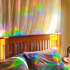May include: A bed with a wooden headboard and two pillows. The pillows and headboard are covered in iridescent rainbow patterns from the sunlight shining through a sheer curtain.