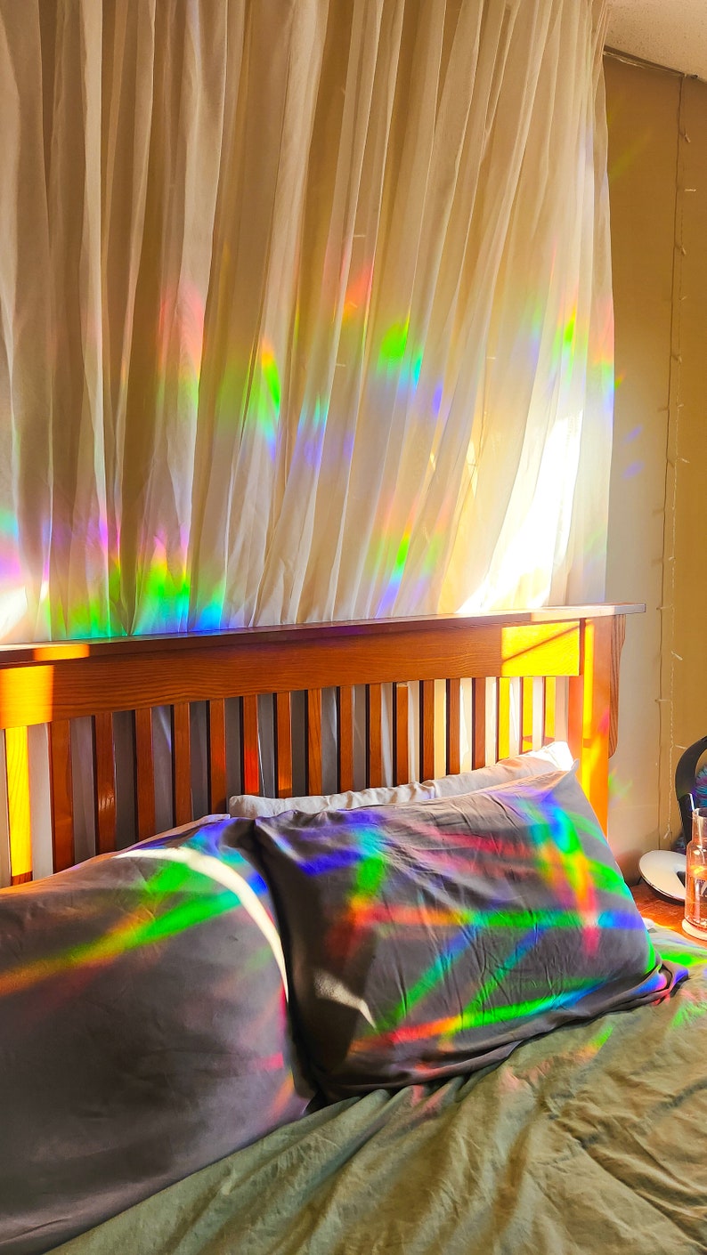 May include: A bed with a wooden headboard and two pillows. The pillows and headboard are covered in iridescent rainbow patterns from the sunlight shining through a sheer curtain.