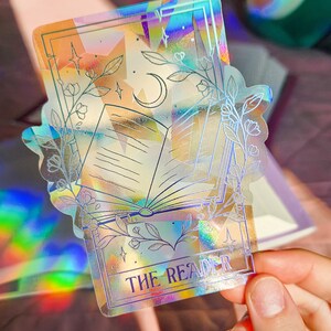 May include: A holographic sticker with a book, moon, and stars. The sticker has a white border and the text "THE READER" at the bottom.