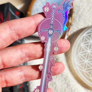 May include: A holographic sticker featuring a sword design with a dragon and stars. The sword has a purple and white color scheme with the words "Mind Cleaver" written on it. The sticker is held up by a hand.