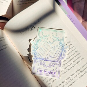May include: A holographic sticker with a purple, blue, and green gradient. The sticker features a line drawing of an open book with a crescent moon and stars above it. The text "THE READER" is printed in purple at the bottom of the sticker.