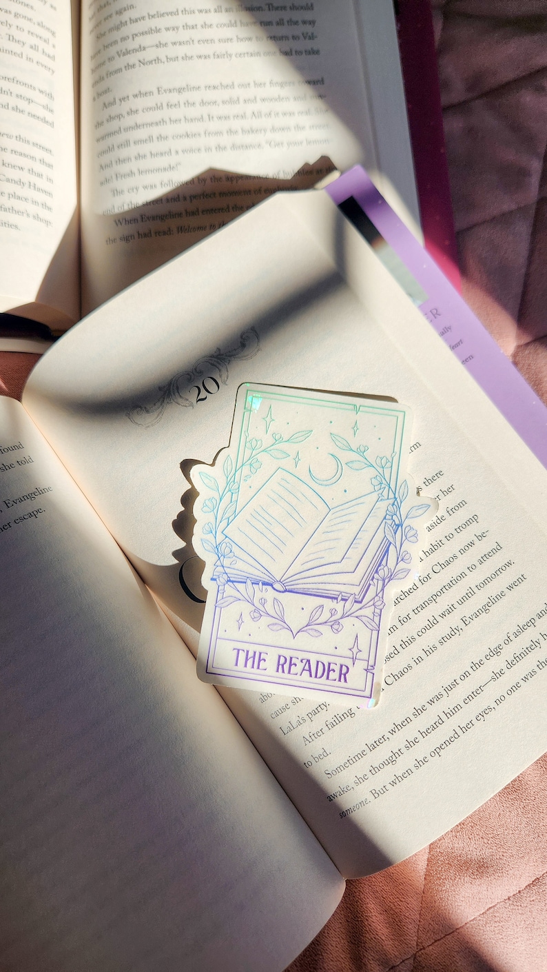 May include: A holographic sticker with a purple, blue, and green gradient. The sticker features a line drawing of an open book with a crescent moon and stars above it. The text "THE READER" is printed in purple at the bottom of the sticker.