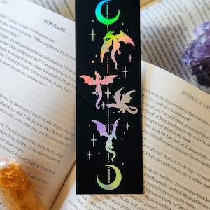 Può includere: Un segnalibro nero con un design olografico di una luna crescente e quattro dragoni con ali arcobaleno iridescenti. I dragoni volano verso l'alto in direzione della luna. Lo sfondo è nero con stelle bianche.