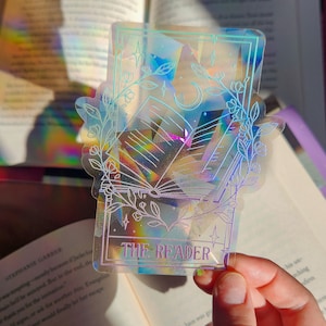 May include: A holographic sticker featuring a book with a crescent moon and stars, surrounded by floral designs. The sticker has a light blue border and the text "THE READER" in purple.