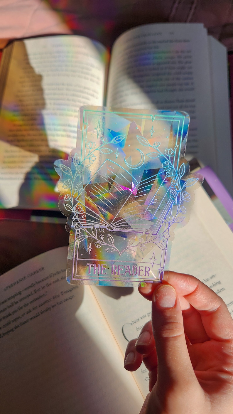 May include: A holographic sticker featuring a book with a crescent moon and stars, surrounded by floral designs. The sticker has a light blue border and the text "THE READER" in purple.