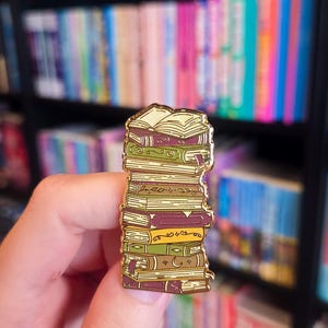 Könnte beinhalten: Ein goldfarbener Emaille-Pin in Form eines Bücherstapels. Die Bücher sind in verschiedenen Farben gehalten, darunter Grün, Braun und Gelb, mit aufwendigen Details. Der Pin wird in einer Hand gehalten, mit einem Bücherregal im Hintergrund.