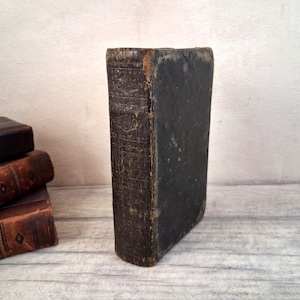 Peut inclure: Un livre ancien sombre et patiné, posé verticalement, doté d'une couverture noire usée et d'un dos orné de motifs décoratifs en relief. À côté, une petite pile de trois livres reliés en cuir brun avec des détails visibles sur le dos.