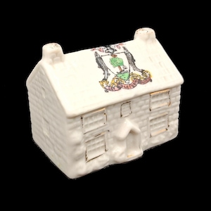 Pode incluir: Uma pequena casa de cerâmica branca com um padrão de tijolos texturizados. O telhado apresenta um desenho de brasão colorido. As janelas e a porta são delineadas em dourado. A casa tem duas chaminés e uma pequena porta.