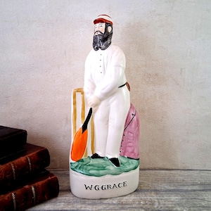 Könnte beinhalten: Keramikfigur eines Mannes in weißer Cricket-Uniform, der einen Schläger hält. Die Figur hat einen schwarzen Bart und eine rote Kappe. Der Sockel ist grün mit dem Text "W.G. GRACE". Eine rosa Jacke ist auf der Rückseite zu sehen.