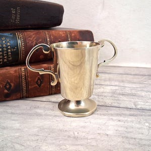 May include: A polished brass trophy cup featuring a smooth, tapered body, a circular pedestal base, and two ornate, curved handles on opposite sides. The metallic surface reflects light, highlighting its golden tone and clean, minimalist design.