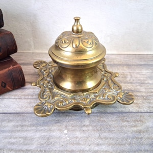 Puede incluir: Tintero antiguo de latón con un diseño floral decorativo. El tintero tiene una tapa abovedada y se asienta sobre una base cuadrada con detalles ornamentados. Este tintero es un artículo vintage, probablemente de principios del siglo XX. Una hermosa pieza de decoración de escritorio.