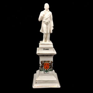 May include: A white ceramic statue of a man in a coat and scarf, standing on a pedestal. The pedestal is inscribed with "Burns" and features a coat of arms with "Aberdeen". The statue is set against a black backdrop.