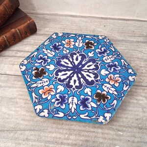 May include: A hexagonal ceramic tile featuring a vibrant turquoise background with a symmetrical floral pattern in shades of dark blue, white, and soft orange. The intricate design radiates from the center, covering the entire surface of the tile.