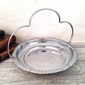 May include: A round silver-toned metal basket featuring a polished interior, a decorative pierced lattice border, and a tall, arched handle with a distinct curved silhouette. The rim of the basket is detailed with a textured, repeating pattern.
