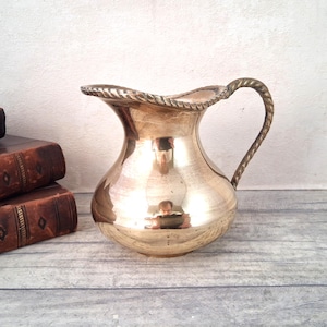 May include: A polished brass pitcher featuring a rounded body, a wide pouring spout, and a decorative twisted rope-style handle. The rim of the pitcher also displays a matching twisted rope texture, creating a cohesive metallic design.