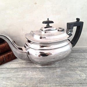 May include: A polished silver-toned teapot featuring a curved spout, a flat lid with a small black knob, and a black angular handle. The teapot has a rounded body and is positioned next to a stack of two brown books with visible text on spines.
