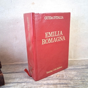 Peut inclure: Un livre rouge intitulé "Emilia Romagna" avec le texte "Guida D'Italia" en haut. Le livre est du "Touring Club Italiano". Un signet rouge est visible.