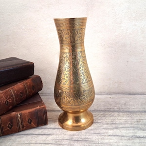 May include: A tall, slender brass vase with a flared rim and a rounded base. The surface of the metal is decorated with intricate, etched floral patterns and horizontal bands. The vase has a polished gold-toned finish with dark, recessed detailing.