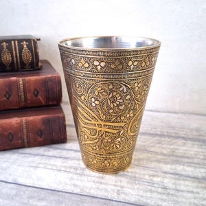 Op de afbeelding: Een decoratieve metalen beker met ingewikkelde bloemen- en geometrische patronen in goud- en zilverkleuren. De beker heeft een uitlopende vorm en wordt waarschijnlijk gebruikt om uit te drinken. De achtergrond bevat antieke boeken.