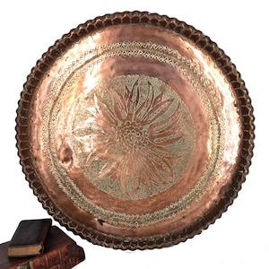 May include: A large, round copper tray featuring a scalloped edge and an intricate, etched floral design in the centre. The metal surface displays a warm, metallic reddish-brown tone with detailed engravings of petals and geometric borders.