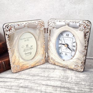 Peut inclure: Un cadre double articulé argenté présentant une ouverture photo ovale à gauche avec le texte Silver Plated et Living Debenhams, et une horloge analogique à droite avec un cadran blanc, des aiguilles noires et des chiffres romains noirs.