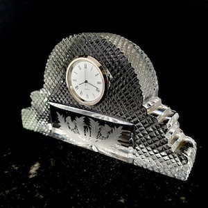 Puede incluir: Un reloj de cristal con una esfera clara y agujas negras. El reloj está colocado sobre una base de cristal con un patrón de diamante y un diseño grabado de palmeras. El reloj es una pieza decorativa para un hogar u oficina.