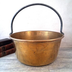 May include: An antique brass cauldron with a dark metal handle. The cauldron has a wide, rounded shape and a slightly textured surface. The handle arches over the top, attached to the sides. The cauldron sits on a wooden surface.