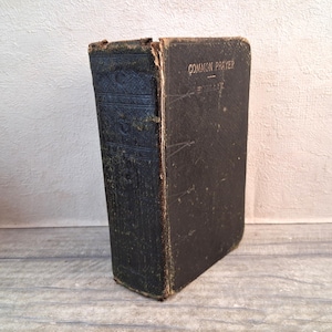 May include: An aged, dark-coloured book with the title "COMMON PRAYER" embossed on the cover. The book shows signs of age, with worn edges and a weathered appearance. The spine has decorative embossed patterns.