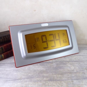 Może przedstawiać: Srebrny i czerwony zegar cyfrowy z dużym, żółtym wyświetlaczem pokazującym godzinę 9:34. Zegar ma czerwoną ramkę i srebrną tarczę.