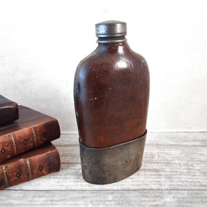 Könnte beinhalten: Eine antike braune Lederflasche mit Metallboden und -kappe. Die Flasche hat eine abgerundete Form und ein abgenutztes Aussehen, was auf Alter und Gebrauch hindeutet. Die Metallkomponenten weisen Oxidationsspuren auf.