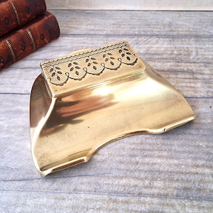 Może przedstawiać: Polerowana mosiężna popielniczka z dekoracyjnym kwiatowym wzorem na górnej krawędzi. Popielniczka ma zakrzywiony kształt i stoi na drewnianej powierzchni. Antyczne książki w tle.