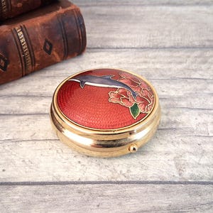 May include: A round gold-tone metal compact case featuring a textured red enamel lid decorated with a blue dolphin illustration and pink floral accents. The circular container has a polished metallic finish and a small side latch for opening.