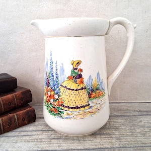 Può includere: Brocca in ceramica color crema con un'illustrazione dettagliata di una donna in abito giallo e scialle verde, con fiori. La brocca ha un manico curvo e un beccuccio. Il design include fiori colorati e un sentiero.