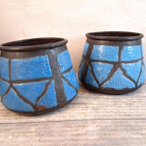 Pode incluir: Dois vasos de cerâmica com um design geométrico azul e castanho. Os vasos têm uma abertura larga e uma forma cónica. As secções azuis têm um padrão texturizado, e as secções castanhas criam um design semelhante a uma grelha. Os vasos estão numa superfície de madeira.
