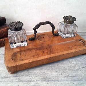 Peut inclure: Un support de bureau en bois avec deux encriers en verre carrés dotés de couvercles en métal ornementés. Une poignée centrale en métal incurvé sépare les encriers, avec une rainure pour stylo sur la base rectangulaire.