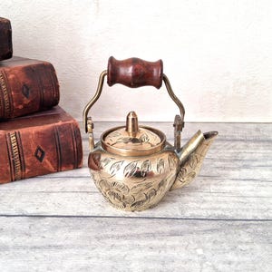 May include: A small brass teapot featuring an intricate floral engraving on the body and lid. The teapot has a hinged metal handle with a dark brown wooden grip and a short, angled spout. The surface of the brass shows a polished metallic finish.