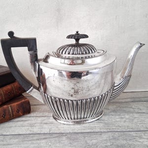 May include: A silver-plated teapot featuring a dark wooden handle and a matching dark wooden finial on the lid. The body of the teapot is decorated with vertical fluted detailing along the lower section and a smooth, reflective upper surface.