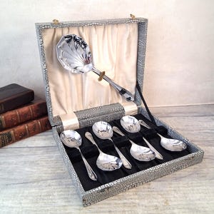Può includere: Un set di cucchiai da portata d'argento e un grande cucchiaio da portata in una scatola di similpelle grigia con fodera in raso color crema. I cucchiai sono disposti su una base di velluto nero. La scatola è aperta.